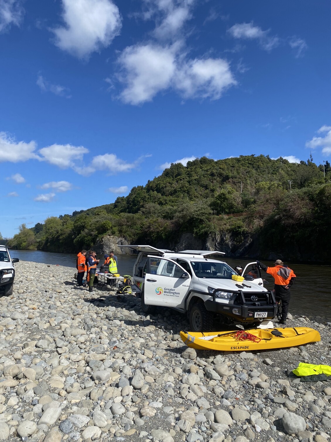Council staff in high visibility clothing prepare monitoring equipment beside Greater Wellington vehicles and a yellow kayak on a stony riverbank under a blue sky.