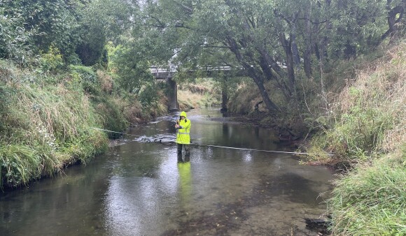 A Greater Wellington field worker in high visibility wet weather gear stands in a shallow stream beneath Palmers Bridge, measuring water levels, with trees lining both banks.